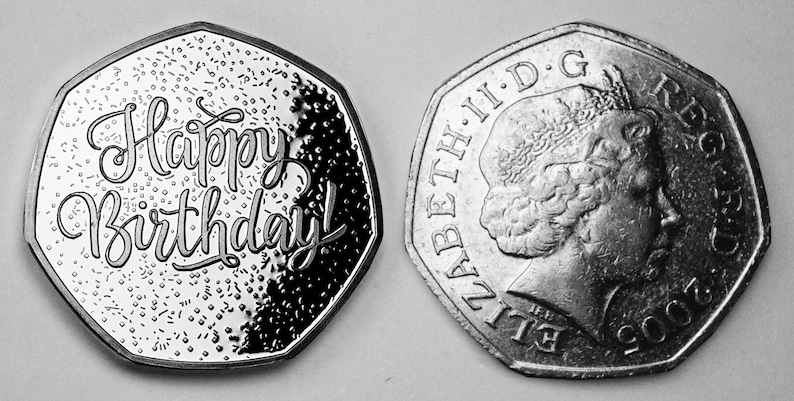 May include: A silver coin with a domed, octagonal shape. One side features the inscription "Happy Birthday!" in a stylized font. The other side features a profile of Queen Elizabeth II and the inscription "ELIZABETH II D.G. REG. F.D. 2002".