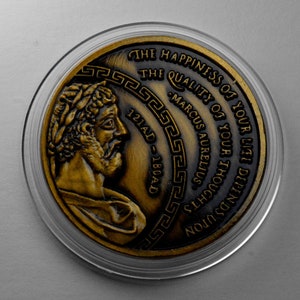 May include: A gold-colored coin with a profile of a man on one side and an inscription that reads "THE HAPPINESS OF YOUR LIFE DEPENDS UPON THE QUALITY OF YOUR THOUGHTS - MARCUS AURELIUS 121AD - 180AD" on the other side.