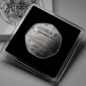 May include: A silver coin with a black and white image of a flying saucer and the text "Area 51 Nevada, USA" on it. The coin is in a clear plastic case with a black background.