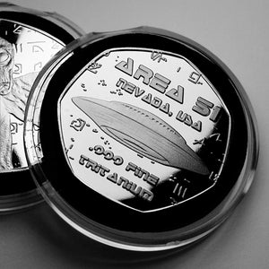 Op de afbeelding: Een zilveren munt met een zwart-wit afbeelding van een UFO en de tekst "AREA 51 NEVADA, USA" en "ONE FINE TRITANIUM" erop.