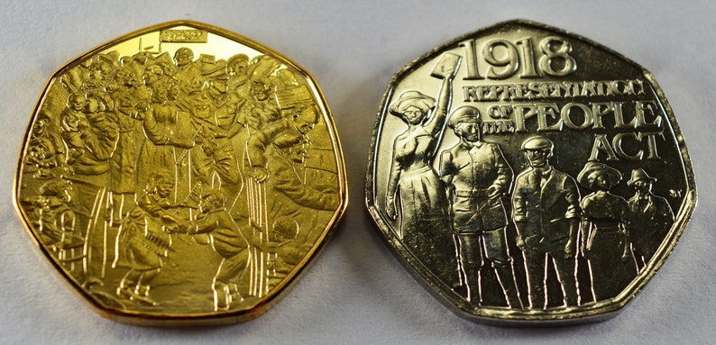 May include: Two gold-colored commemorative coins depicting the 1918 Representation of the People Act. The obverse side shows a group of people, while the reverse side shows a group of people standing with the text "1918 REPRESENTATION OF THE PEOPLE ACT".
