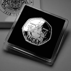 Puede incluir: Una moneda de plata con una imagen en blanco y negro de un Buda riendo. La moneda está en una caja de plástico transparente. La moneda dice "Laughing Buddha" y "Lucky".