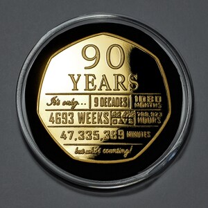 May include: A gold-coloured coin with a black background and the inscription "90 YEARS" in large letters. The coin also includes the text "It's only... 9 DECADES | 1080 MONTHS | 4693 WEEKS | 32,872 DAYS | 788,923 HOURS | 47,335,389 MINUTES but who's counting!"