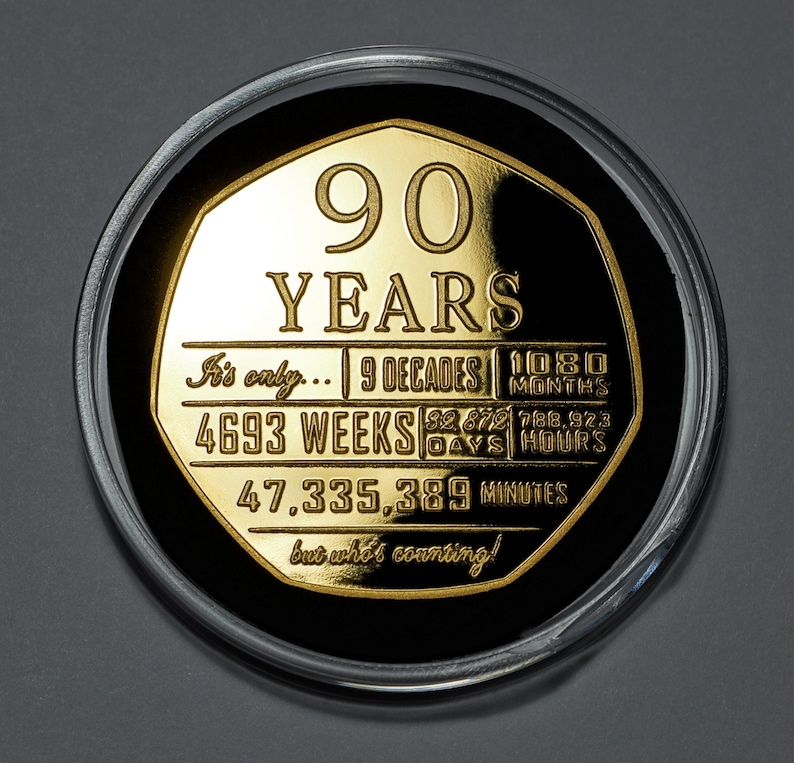 May include: A gold-coloured coin with a black background and the inscription "90 YEARS" in large letters. The coin also includes the text "It's only... 9 DECADES | 1080 MONTHS | 4693 WEEKS | 32,872 DAYS | 788,923 HOURS | 47,335,389 MINUTES but who's counting!"