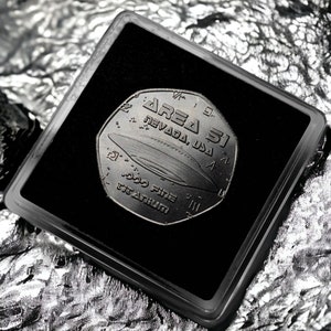 May include: A black and silver coin with the words "Area 51 Nevada, USA" and "999 Fine Titanium" inscribed on it. The coin features a UFO design.