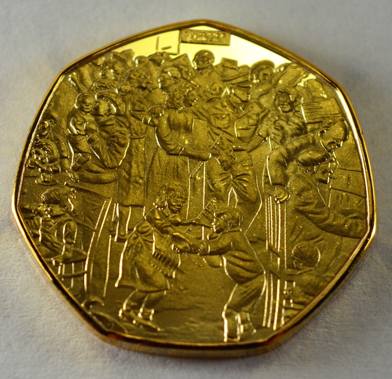 May include: A gold-colored commemorative coin featuring a detailed relief design of a crowd of people, including children, gathered together. The coin has a dodecagonal shape and the inscription "2000" is visible on the top.