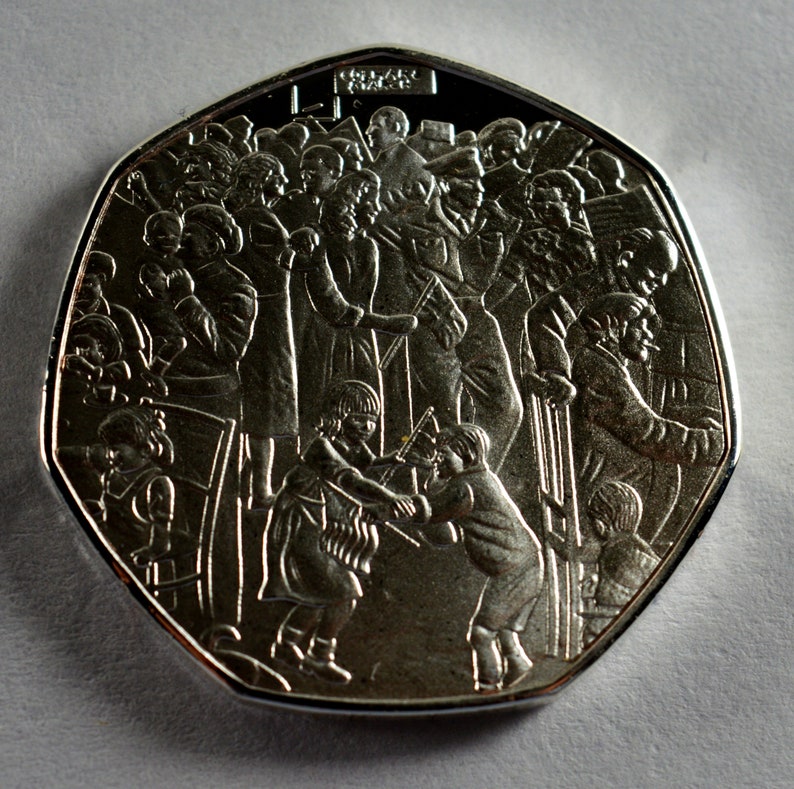 May include: A silver commemorative coin featuring a detailed design of a crowd of people, including children, with the words "One Pound" inscribed on the top.