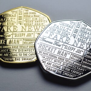 May include: Two commemorative coins, one gold-toned and one silver-toned, with engraved text. The coins feature phrases like "FAKE NEWS," "ROCKET MAN," and "DRAIN THE SWAMP." The design includes multiple lines of text.