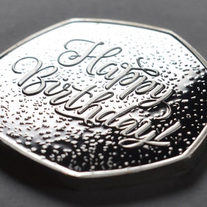 以下が含まれることがあります： 「Happy Birthday」の文字が刻まれた銀色のコイン。コインはテクスチャ加工された表面と光沢のある仕上げが施されています。コインは濃いグレーの背景に置かれています。