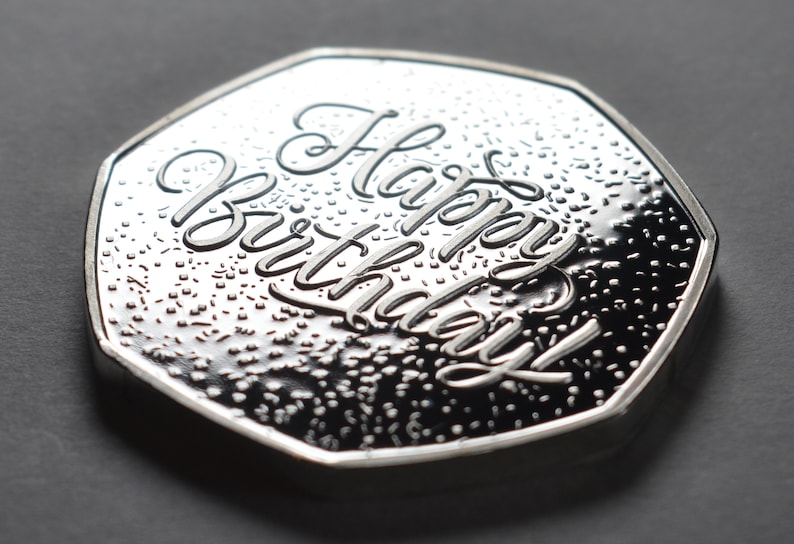 以下が含まれることがあります： 「Happy Birthday」の文字が刻まれた銀色のコイン。コインはテクスチャ加工された表面と光沢のある仕上げが施されています。コインは濃いグレーの背景に置かれています。