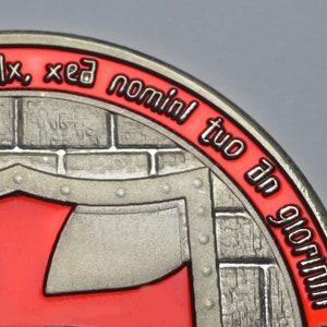 Op de afbeelding: Een zilverkleurige metalen munt met een rood en zwart ontwerp. De munt heeft een bakstenen muur met een rood schild en de tekst "dulx, xed nominl tuo dn glorium".