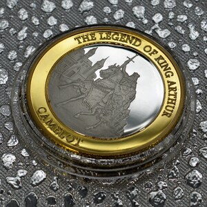 May include: A silver coin with a gold rim featuring a depiction of King Arthur on horseback, with the text "The Legend of King Arthur Camelot" around the edge.