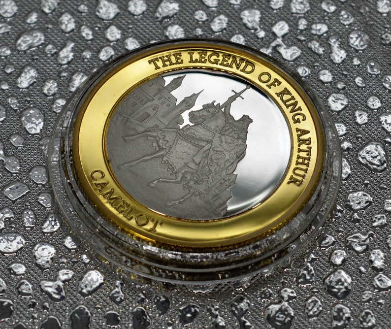 May include: A silver coin with a gold rim featuring a depiction of King Arthur on horseback, with the text "The Legend of King Arthur Camelot" around the edge.