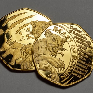 Könnte beinhalten: Zwei goldfarbene Münzen mit einem Design eines mythischen Tieres. Die Münze rechts trägt den Text "BEAST OF BODMIN" und eine Beschreibung des Tieres. Die Münze links zeigt ein Tier mit Krallen und ein kleineres Tier.