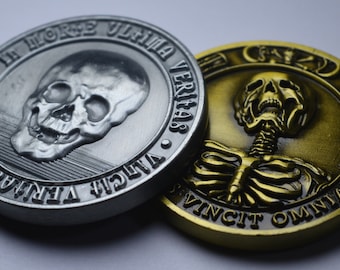 May include: Two detailed metal coins. One is silver with a skull and the inscription "MORS ULTIMA VERITAS". The other is gold-colored, featuring a skeleton and the inscription "OMNIA VINCIT".