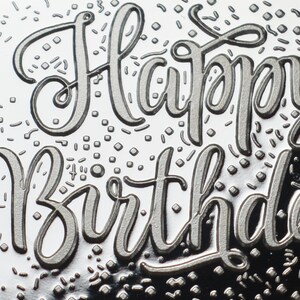 以下が含まれることがあります： 筆記体で「Happy Birthday」の文字がエンボス加工された、銀色のデザインのクローズアップ。デザインには、小さく散らばった紙吹雪の形が含まれており、お祝いの雰囲気を醸し出しています。