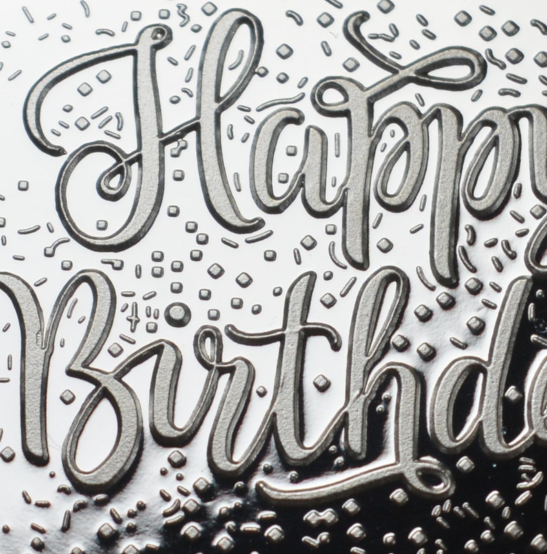以下が含まれることがあります： 筆記体で「Happy Birthday」の文字がエンボス加工された、銀色のデザインのクローズアップ。デザインには、小さく散らばった紙吹雪の形が含まれており、お祝いの雰囲気を醸し出しています。