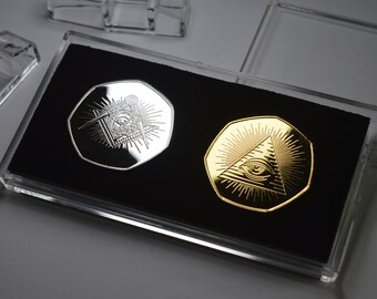 Puede incluir: Dos monedas de plata y oro con un diseño de ojo que todo lo ve en una vitrina de plástico transparente. Las monedas son octogonales y presentan una pirámide con un ojo en la parte superior.