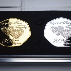 Puede incluir: Dos monedas conmemorativas en una caja de terciopelo negro. Las monedas son de oro y plata y presentan un diseño de dos corazones entrelazados con el texto "For Your Pearl Wedding Anniversary" y "30 Years Together".