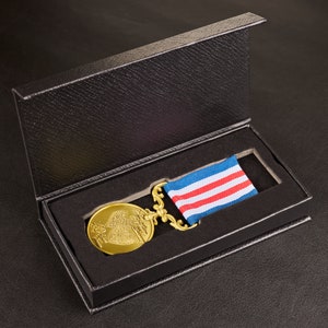 May include: A gold medal with a heart design and a red, white and blue ribbon is displayed in a black velvet box.