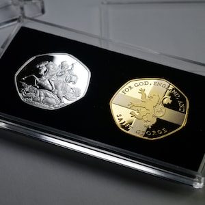 Puede incluir: Dos monedas conmemorativas de plata y oro en una vitrina de plástico transparente. La moneda de plata muestra una representación de San Jorge matando a un dragón. La moneda de oro muestra un león y el texto "For God, England and Saint George".