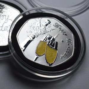 May include: A silver coin featuring a design of two champagne flutes and a bottle of champagne with the text "Happy Birthday" around the edge.