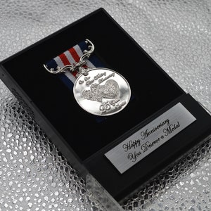 May include: Silver medal with a red, white, and blue ribbon in a black box. The medal features a heart with wings and the text "Our 25th Wedding Anniversary". The box has a silver plaque with the text "Happy Anniversary You Deserve a Medal".