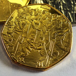 May include: A gold-colored coin with a raised design of people skiing. The coin has a rounded, octagonal shape.