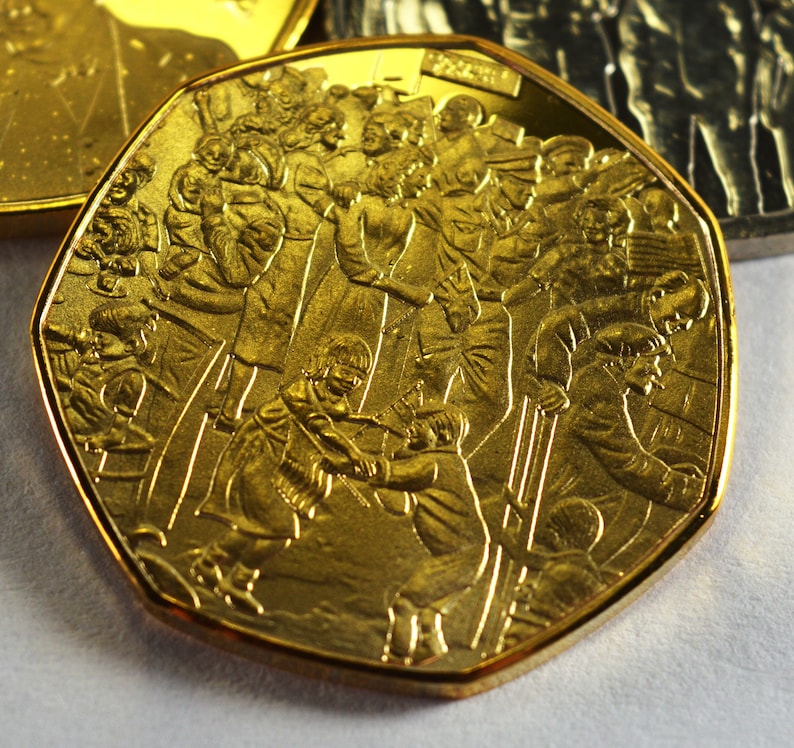 May include: A gold-colored coin with a raised design of people skiing. The coin has a rounded, octagonal shape.