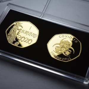 以下が含まれることがあります： 透明なプラスチック製のディスプレイケースに入った、金色の記念硬貨2枚。1枚目の硬貨にはペスト医師が描かれ、「I Survived 2020」の文字が刻まれています。2枚目の硬貨にはガスマスクが描かれ、「COVID-19 Coronavirus」の文字が刻まれています。