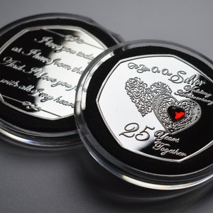 May include: Two silver-toned coins in clear plastic cases. The coin on the left has an inscription that reads "I love you today as I have from the first. And I love you now with all of my heart". The coin on the right has a red heart with a silver filigree design and the inscription "For You On Our Silver Wedding Anniversary 25 Years Together".