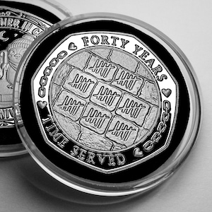 May include: A silver-toned commemorative coin encased in a clear protective capsule. The coin features a central grid of nine tally mark squares, surrounded by the text "FORTY YEARS" and "TIME SERVED," accented with small hearts and chain links.