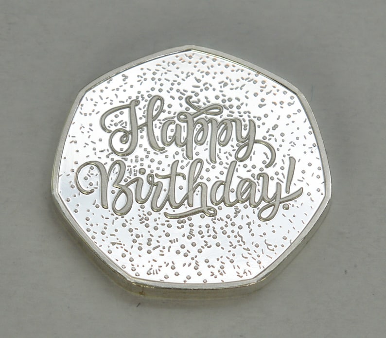 May include: Silver coin with a textured surface and the words "Happy Birthday!" inscribed in a cursive font.