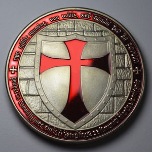 Op de afbeelding: Een zilveren munt met een rood kruis op een schild, omringd door een bakstenen muurontwerp. De munt heeft een Latijnse tekst rond de rand, waaronder "commilijunes cruisu templique salomunici nicrusalemitanis".