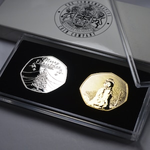 Puede incluir: Dos monedas conmemorativas de plata y oro en una caja de plástico transparente. La moneda de plata presenta un árbol de Navidad y las palabras "Merry Christmas". La moneda de oro presenta un muñeco de nieve.