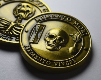 May include: Two gold-colored metal coins with Latin inscriptions. The first coin features a skeleton with the inscription "Memento Mori". The second coin features a skull with an hourglass, a flower, and the inscription "Memento Mori Memento Vivere".