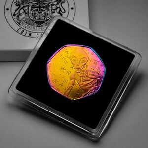 May include: A gold-colored coin with a holographic finish features an alien with large eyes and a detailed design. The coin is in a black display case.