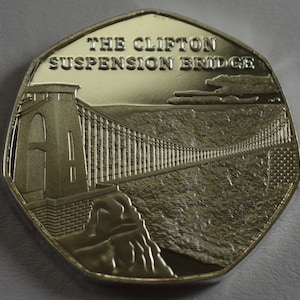 May include: A silver-colored commemorative coin featuring a detailed image of the Clifton Suspension Bridge in Bristol, England. The coin has the words "The Clifton Suspension Bridge" inscribed on the top.