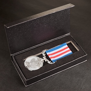 May include: A silver medal with a heart design and a red, white and blue ribbon is displayed in a black velvet box.