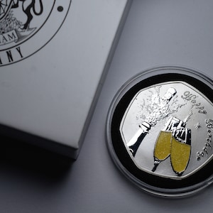May include: A silver coin featuring a design of a champagne bottle and two glasses filled with a gold liquid. The coin is inscribed with the words "Happy Birthday".