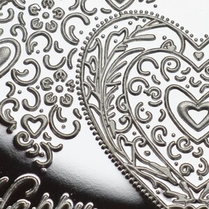 May include: A silver heart-shaped ornament with an intricate embossed design. The design features a floral pattern with swirls and dots.