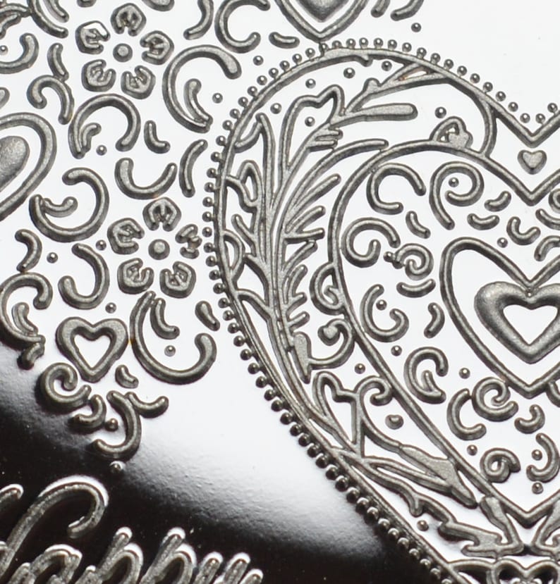May include: A silver heart-shaped ornament with an intricate embossed design. The design features a floral pattern with swirls and dots.