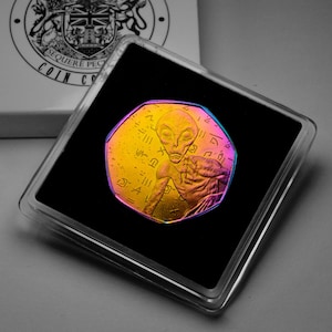 May include: A colorful coin featuring an alien figure with hieroglyphic-style symbols. The coin is encased in a clear plastic holder on a black background. The coin's colors shift from gold to purple and blue.