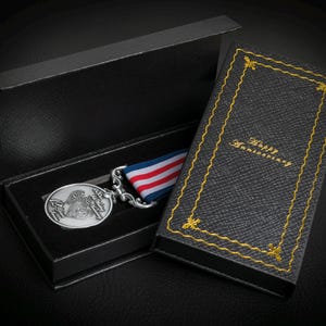 May include: A silver medal with a blue, white, and red ribbon, displayed in an open black presentation box. The box next to it has the words "Happy Anniversary" in gold lettering. The box is black with gold trim.