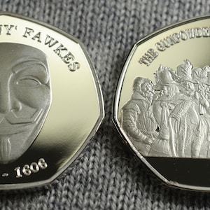 Op de afbeelding: Twee zilveren munten met een verhoogde rand. De voorzijde van de munt toont een zilveren masker met een glimlach en de tekst "GUIDO 'GUY' FAWKES 1570 - 1606". De achterzijde van de munt toont een groep mensen in historische kleding met de tekst "THE GUNPOWDER CONSPIRATORS".
