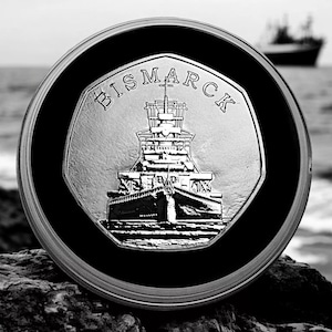 May include: A silver coin with a black rim features a detailed image of the German battleship Bismarck. The coin is inscribed with the word "BISMARCK" around the edge.