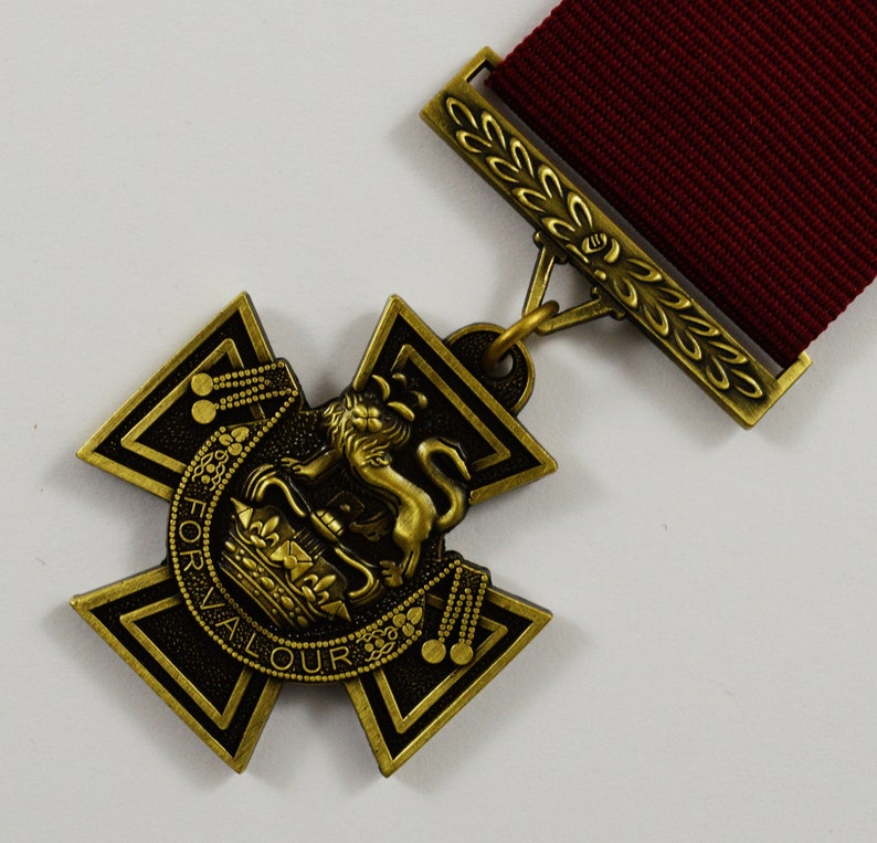 Pu&ograve; includere: Una medaglia dorata con un nastro rosso. La medaglia presenta una croce con un leone e una corona al centro. L'iscrizione "FOR VALOUR" &egrave; visibile sulla medaglia.