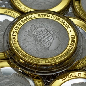 Puede incluir: Una moneda de color dorado con un centro plateado que presenta una huella y el texto "That's one small step for man. One giant leap for mankind."