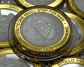 May include: A gold-coloured coin with a silver centre featuring a footprint and the text "That's one small step for man. One giant leap for mankind."