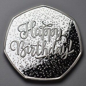 以下が含まれることがあります： 「Happy Birthday!」と書かれた銀色のコイン。コインは独特の多角形の形状をしており、小さな散らばったディテールを持つテクスチャード加工の背景があります。デザインは鮮明で詳細です。